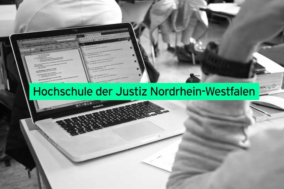 Auf dem Bild ist ein Laptop auf einem Tisch abgebildet und eine Person, die in einem Klassenzimmer davor sitzt. 
