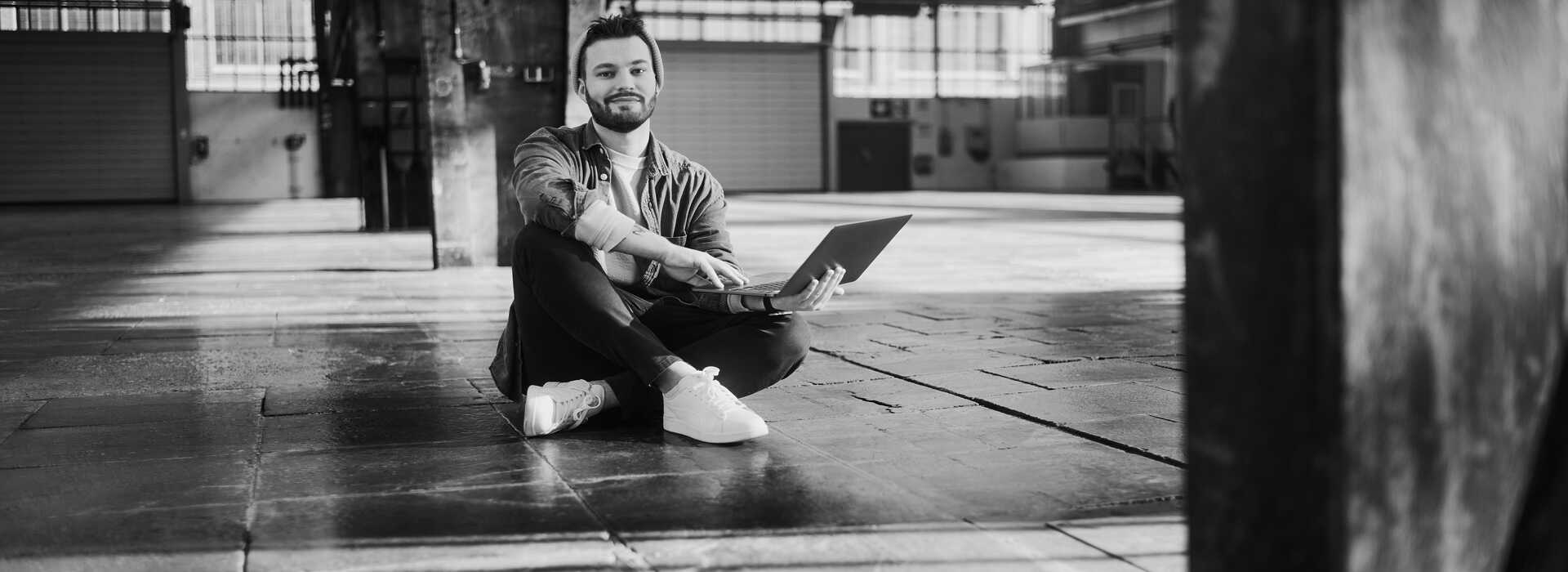 Ein Mitarbeiter sitzt mit aufgeklapptem Laptop auf dem Boden einer leeren Lagerhalle.