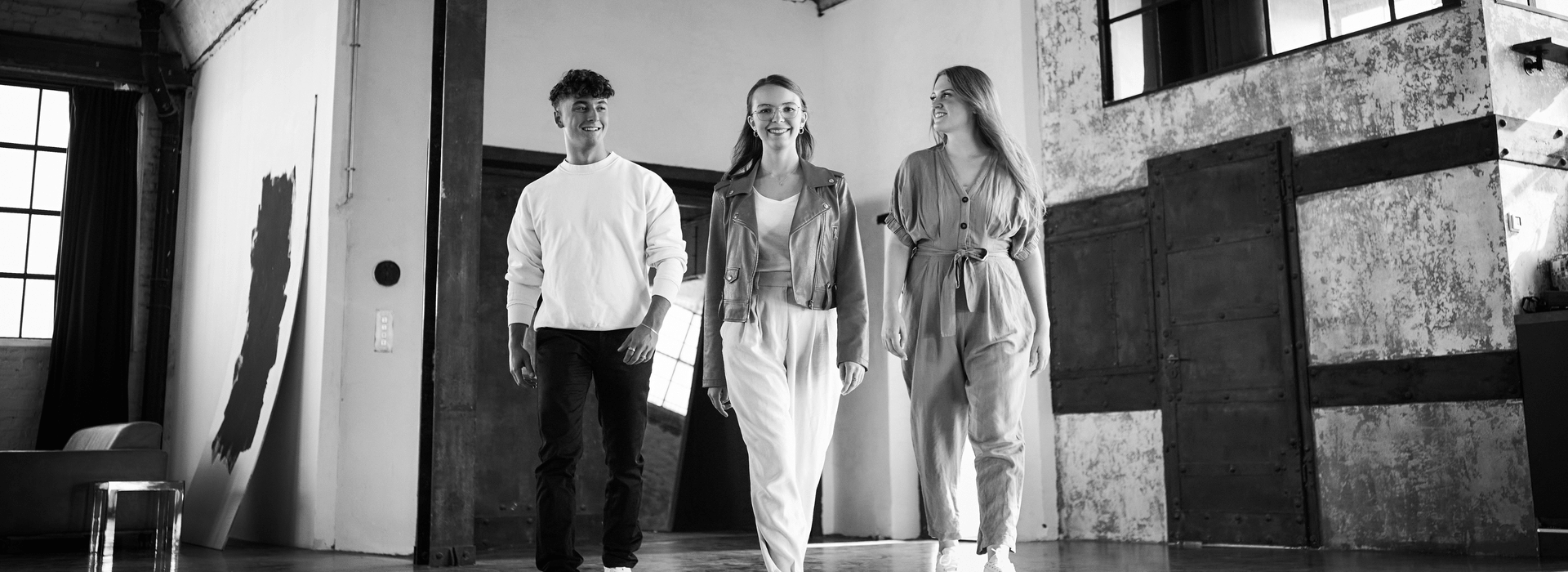 Auf dem schwarz-weiß Bild sind zwei junge Frauen und ein junger Mann abgebildet, die nebeneinander laufen.