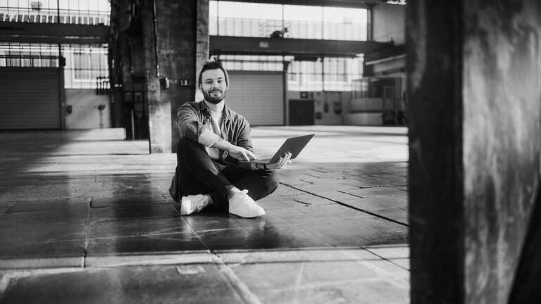 Ein Mitarbeiter sitzt mit aufgeklapptem Laptop auf dem Boden einer leeren Lagerhalle.